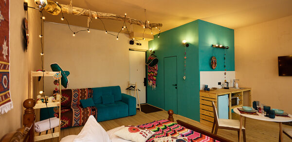 ⁠Лофт в Мексиканском стиле для костюмированных вечеринок. Гостиная с кроватью, диваном, столом и стульями. На голубой стене декор и полка с посудой. Деревянная мебель, яркие ткани с узорами. Верхнее и гирляндное освещение. Деревянный пол, стены светлые. Бежевые и голубые цвета. Телевизор на стене.