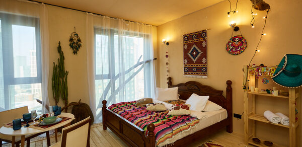 ⁠Лофт в Мексиканском стиле для костюмированных вечеринок. Просторная комната с деревянным полом, большая кровать с текстилем ярких цветов, рядом деревянный стол и два стула. На стене тканевые декорации, полка с декором. Окна в пол, через которые проникает свет. Потолочный декор с лампами, светлый текстиль на окнах.
