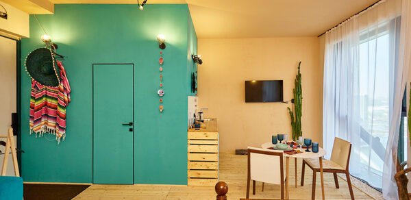 ⁠Лофт в Мексиканском стиле для костюмированных вечеринок. Комната с деревянным полом, бирюзовой стеной и декором. Белые и деревянные стулья вокруг стола с посудой. Потолочные светильники, телевизор на стене, зелёное растение у окна с белыми шторами. Цветные пледы на стуле и стене.