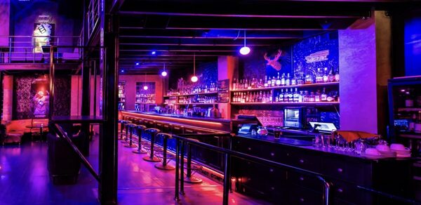 Castle Concert Hall(Концертный зал Замок). Барное пространство с длинной стойкой и высокими стульями, освещение фиолетового и розового оттенков. На заднем плане стеллажи с бутылками, неоновые элементы на стенах. Металлические и деревянные элементы, открытые трубы и балки.