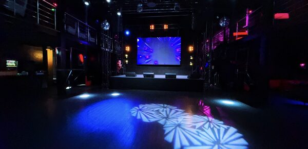 Castle Concert Hall(Концертный зал Замок). Ночной клуб или концертный зал с темным интерьером, большой экран на сцене, разноцветные прожекторы и световые эффекты на полу, металлические перила, второй ярус, черный пол и стены, современные осветительные приборы, отсутствуют посетители.