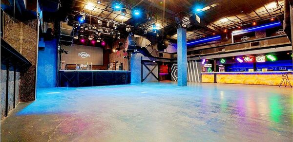 PRAVDA(Правда). Просторный зал на 800 гостей. Широкое внутреннее пространство с бетонным полом. Площадка с оборудованием для выступлений. Стойка бара с подсветкой. Освещение разноцветное, включая прожекторы и неон. Высокий потолок с открытыми металлическими конструкциями. Тёмные стены.