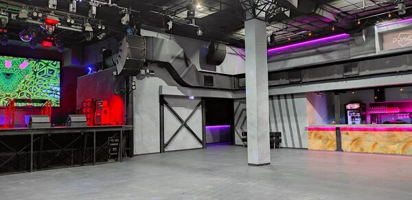 PRAVDA(Правда). Просторный зал на 800 гостей. Просторное помещение с бетонным полом и колонной в центре. Серые стены с черными элементами. Площадка с оборудованием и красной подсветкой. Барная стойка с фиолетовой и розовой подсветкой. Потолочные светильники и аудиосистема.
