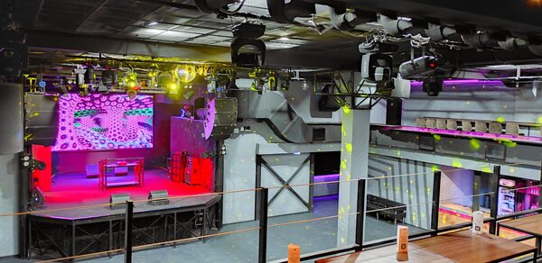 PRAVDA(Правда). Просторный зал на 800 гостей. Клубное помещение с большой сценой, оснащённой экранами и звуковым оборудованием. Верхний этаж с балюстрадой и зонами для сидения. Освещение разноцветное, включая прожекторы. Стены серые, полы тёмные. Потолок в чёрных конструкциях. Отсутствие людей.