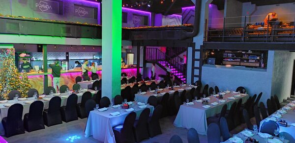 PRAVDA(Правда). Просторный зал на 800 гостей. Большое помещение с несколькими уровнями, длинные банкетные столы с белыми скатертями и черными стульями, яркое цветное освещение, лестница, барная стойка с подсветкой. Стены и потолок темные, оформлены неоновой подсветкой. Текст: "PANDA".