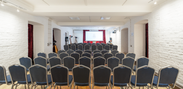 Светлый Лофт. Конференц-зал с белыми кирпичными стенами и деревянным полом. Ряды серых стульев направлены к экрану и проектору. Потолок с белыми панелями и встроенными светильниками. На экране проекция черно-белого логотипа.