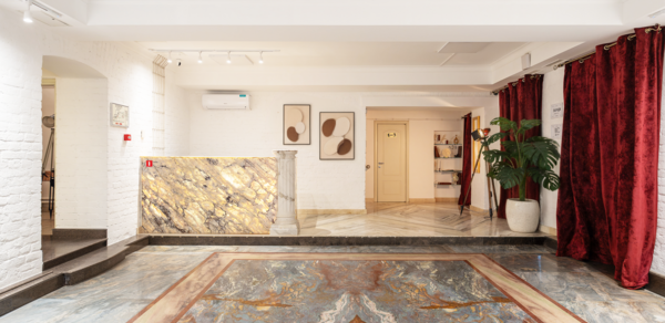 Светлый Лофт. Просторное помещение с мраморным полом и зоной рецепции. Белые кирпичные стены, светильники на потолке. Декоративные панели на стене, красные шторы справа, декоративное растение в горшке, кондиционер, полка с книгами.