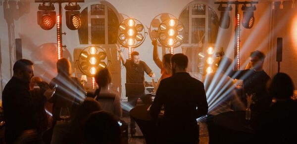 Светлый Лофт. Пространство для вечеринки с диджеем, круглыми высокими столами, людьми, световыми установками и эффектами. Белые кирпичные стены, большие окна, оранжевое освещение, черные мебельные чехлы.