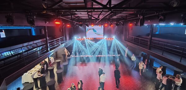 СВОБОДА. Зал на 1200 человек. Многоуровневое пространство для мероприятий с паркетным полом и металлическими перилами. Несколько столов с белыми скатертями и барными стульями. Яркое освещение с прожекторами, сцена с экраном. Люди в зале.