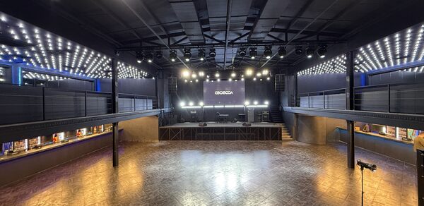 СВОБОДА. Зал на 1200 человек. Просторное темное помещение с паркетным полом, центральной сценой и балконами по обе стороны. Разнообразное освещение, включая потолочные светильники и светодиодные полосы. На сцене черная стена с надписью. Справа микрофон на стойке.