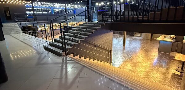 Концертный зал СВОБОДА с танцполом. Многоуровневое пространство с современной лестницей и перилами, плиточный пол, освещение прожекторами, стулья на верхнем уровне, столы на первом этаже, яркие потолочные лампы, темные стены, полированный светлый пол и потолок со светодиодными лампами.