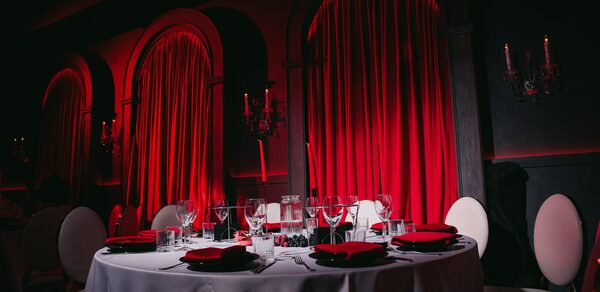 QUQLA. Круглый стол с белой скатертью, сервирован черной и красной посудой; бокалы и столовые приборы. Белые стулья с округлыми спинками. Красные шторы, подсвеченные снизу. Темные стены с арочными нишами и канделябрами.