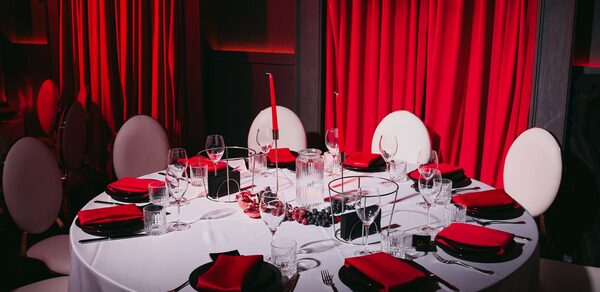 QUQLA. Круглый стол с белой скатертью, сервированный на шесть персон. Красные салфетки, черные тарелки, вилки, ножи, бокалы, свечи. Белые овальные стулья, красные шторы, приглушенное освещение. Декор из ягод.