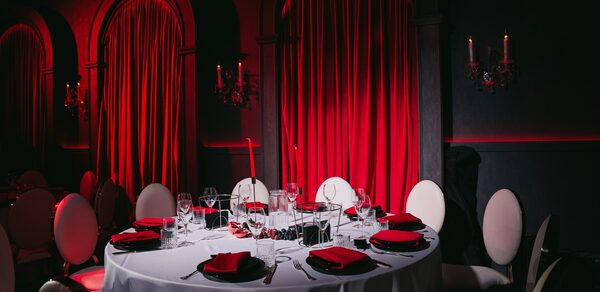 QUQLA. Банкетный зал с круглым столом, сервированным бокалами, приборами и черными тарелками с красными салфетками. Окружен белыми стульями. На стенах красные занавески и подсвечники. Освещение теплое, приглушенное. Цвета: красный, черный, белый.