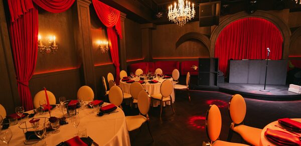 QUQLA. Банкетный зал с круглыми столами, накрытыми белыми скатертями. Красные занавески и декорации, бежевые стулья с подушками. Люстры и бра с теплым освещением. Сцена с микрофоном и звуковой аппаратурой. Тёмный пол, стены в тёмных тонах.