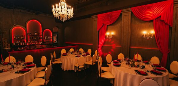 QUQLA. Помещение с круглыми столами, сервированными красно-черными тарелками и бокалами. Светлые стулья, барная стойка с бутылками на полках. Красные драпировки и подсветка, люстры и настенные светильники. Темные стены, арочные ниши за баром.