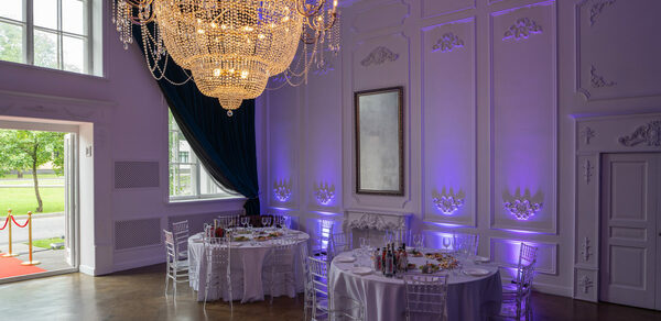 Лофт КОСМОС с летней верандой. Большая комната с роскошной люстрой, двумя круглыми столами с белыми скатертями и прозрачными стульями. Декоративные панели на стенах, одно зеркало. Фиолетовая подсветка, темно-синие шторы, деревянный пол, высокие окна и двери.