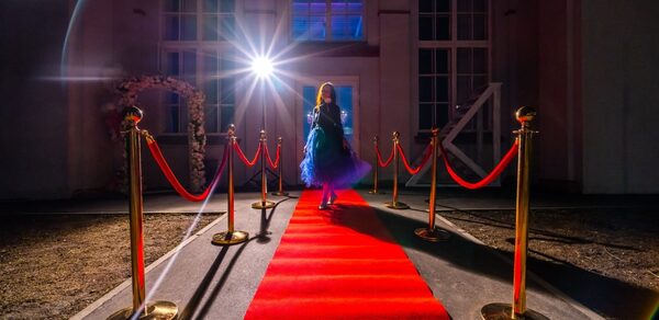 Лофт КОСМОС с летней верандой. Красная ковровая дорожка окружена бархатными канатами на золотых стойках. Пространство перед зданием с высокими окнами. Яркий свет направлен на дорожку. Декоративная арка справа. Северное сияние в небе.