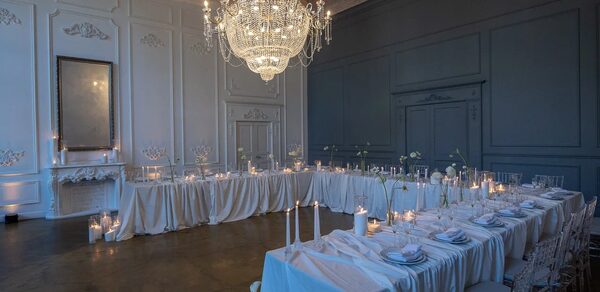 Лофт КОСМОС с летней верандой. Праздничное пространство с длинными столами в форме буквы «Г», покрытыми белыми скатертями и сервировочными наборами. Стулья прозрачные, вокруг свечи. Центральный элемент — большой хрустальный светильник. Стены белого и серого цветов с панелями.