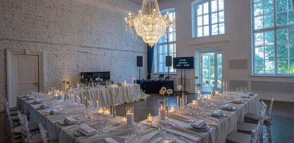 Лофт КОСМОС с летней верандой. Пространство для мероприятий с белыми кирпичными стенами, большим люстрой, светлыми скатертями, сервировкой столов, прозрачными стульями. Высокие окна, круглые и квадратные столы, свечи. Освещение теплое, дневной свет.