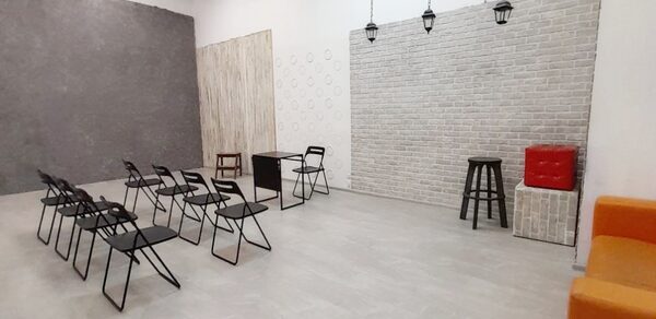 Интер-Фото. Просторное помещение с кирпичной и бетонной стенами, расположенными под углом. На полу светлая плитка. В центре черные складные стулья в два ряда с небольшим столом. Подвесные светильники. По краям стула, стол, пуф и барный стул.