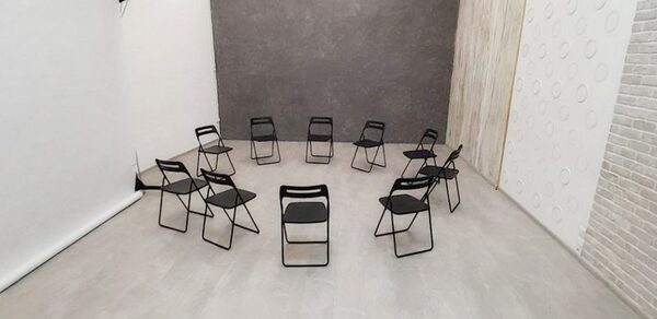 Интер-Фото. Просторное помещение с белыми стенами и светлым плиточным полом. В центре расставлены черные складные стулья по кругу. Стены декорированы серыми и белыми текстурными панелями. Есть рулон белого фона, установленного на стойке.