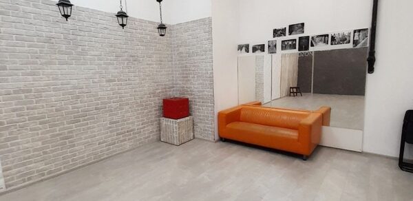 Интер-Фото. Просторная комната с белым кирпичным декором стен. На полу светлая плитка. Оранжевый диван расположен перед зеркальной стеной. Подвесные черные светильники. На стене черно-белые фотографии. Белая квадратная корзина и красный куб.