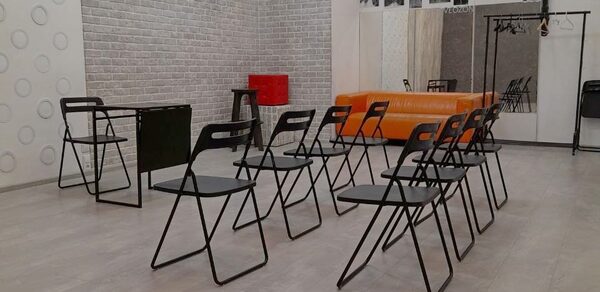 Интер-Фото. Просторное помещение с паркетным полом, кирпичными стенами и белым потолком. В центре расположены ряды черных складных стульев, оранжевый диван у стены, черный стол, на стене фотографии и надпись "WELCOME". Освещение естественное.