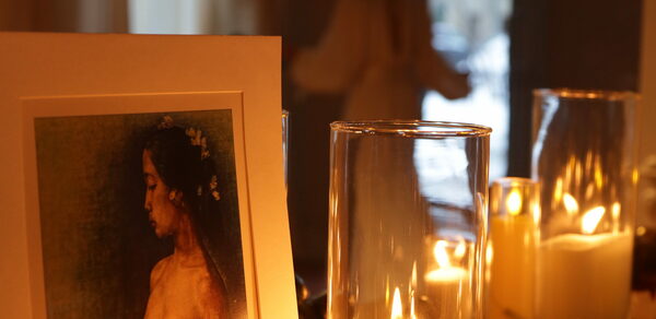 Лофт Поле. Уютный зал для мероприятий. Крупный план свечей в прозрачных стеклянных подсвечниках. На переднем плане изображение с портретом женщины. Вдалеке размытый силуэт человека у окна. Интерьер в тёплых тонах.