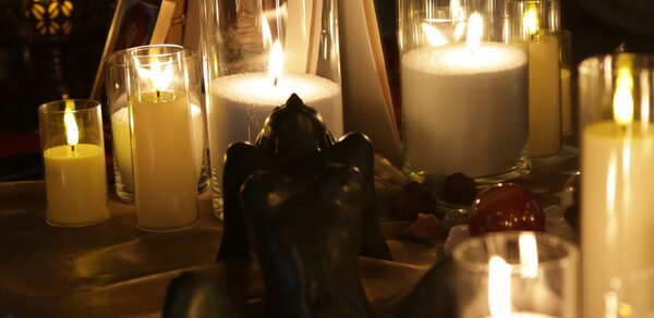 Лофт Поле. Уютный зал для мероприятий. Свечи в прозрачных стаканах и большие белые свечи на столе, украшенном тканью. Темная фигурка декора, вокруг небольшие камни. Освещение теплое и мягкое, создающее атмосферу уюта и интимности.