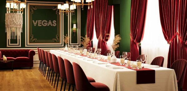 Вегас. Атмосферный лофт с акцентным дизайном. Банкетный зал с длинным столом, накрытым белой скатертью и красными салфетками, окружённый бордовыми стульями. Красные бархатные шторы и зелёные стены. Светильники с подсвечниками. На стене текст: "VEGAS". Пол из светлого дерева.
