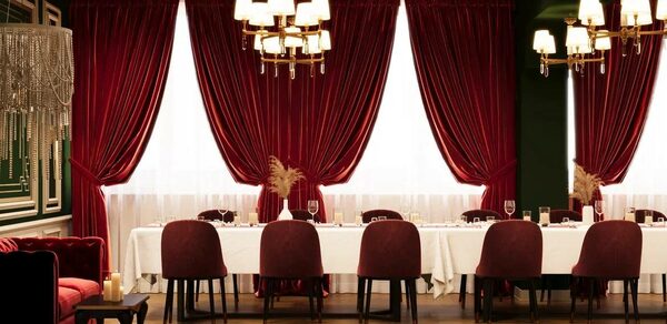 Вегас. Атмосферный лофт с акцентным дизайном. Банкетный зал с длинным столом, накрытым белой скатертью, окружённый бордовыми стульями. Красные бархатные шторы, зелёные стены, деревянный пол. Потолочные и настенные светильники с белыми абажурами. Декор из сухих растений, свеча на кофейном столике.