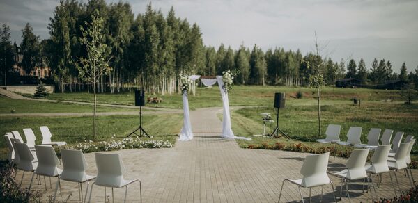 Эко-отель "Джао Да!Ча". Шатер-Сфера. Открытое пространство с кирпичной дорожкой, окружённой газоном. Арка, украшенная белым текстилем и цветами. По бокам арки установлены колонки на стойках. По обе стороны дорожки расположены белые стулья в два ряда, образуя полукруг. Позади деревья.