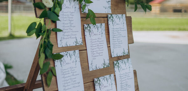 Эко-отель "Джао Да!Ча". Шатер-Сфера. Деревянная подставка с планом рассадки гостей оформлена белыми карточками с текстом и украшена зелеными листьями и цветами. На заднем плане виден участок земли с зелёной травой и зданием.