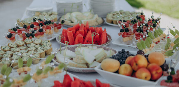 Эко-отель "Джао Да!Ча". Шатер-Сфера. Стол с тарелками, заполненными фруктами, такими как арбузы, дыня, абрикосы и виноград. Также представлены десерты и закуски на шпажках. Посуда белая, вокруг видна зелёная трава и расставлены стаканы.