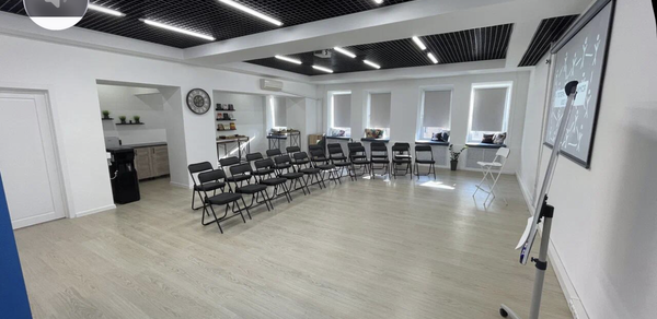 Уютный тренинговый зал на Таганской. Просторное помещение в светлых тонах, с деревянным полом, белыми стенами и потолками с черными решетками и встроенным освещением. Ряды складных стульев, магнитно-маркерная доска на стенде, кухня с посудой слева, окна с жалюзи.
