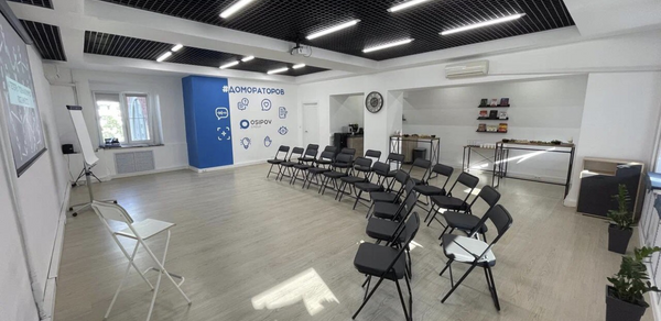 Уютный тренинговый зал на Таганской. Просторное помещение с белыми стенами и черным потолком, оборудовано рядами складных стульев, проектором и экраном, мольбертом. Стена с синим акцентом и иконками. В углу столы с закусками и напитками. Панорамное окно, элементы озеленения.