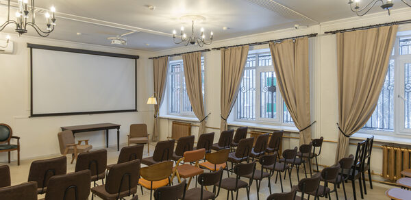 Open Space (Открытое пространство) для мероприятий. Конференц-зал с рядами стульев, деревянными полами и белыми стенами. Экран для проекции, столы и кресла. Светильники на потолке и торшер. Большие окна с бежевыми шторами. Металлические решетки на окнах, светлое освещение.