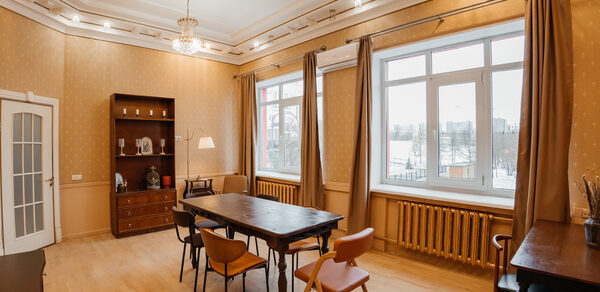 Open Space (Открытое пространство) для мероприятий. Просторная комната с деревянным полом, светлыми стенами и высоким потолком с декоративной лепниной. Центральное освещение люстрой, дополнительные светильники по периметру. Большие окна с бежевыми шторами, деревянный стол, стулья, шкаф с посудой, кресло.