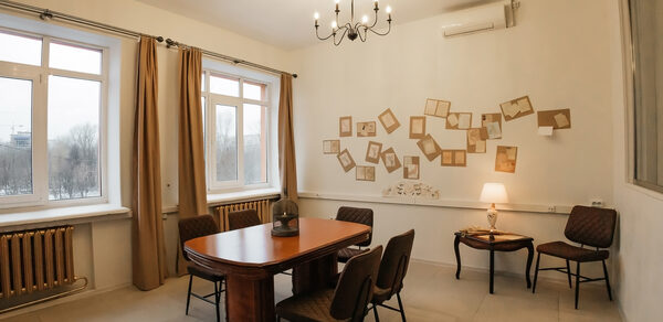 Open Space (Открытое пространство) для мероприятий. Комната с прямоугольным столом из дерева и четырьмя стульями, освещенная подвесной люстрой и настольной лампой. Два окна с занавесками, стена украшена рамками. На полу плитка, в углу стул и столик, белые стены, кондиционер.