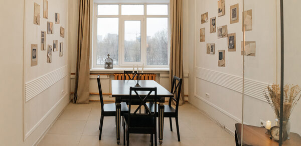 Open Space (Открытое пространство) для мероприятий. Узкая комната с белыми стенами, большая центральная оконная рама, задернутая бежевыми шторами. Темный прямоугольный стол с четырьмя стульями по бокам. На стенах развешаны фото и рисунки. Светлые плиточные полы, зеркало справа, деревянная тумбочка.