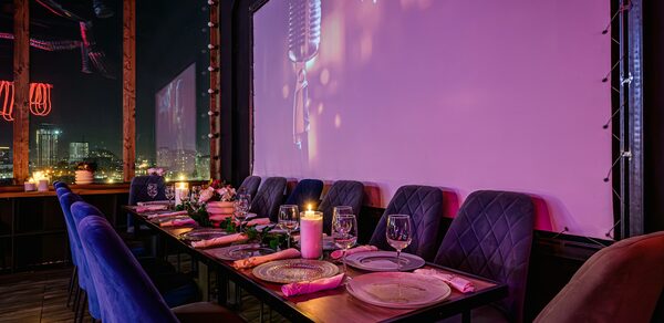 Лофт Овертон. Уютное помещение с длинным деревянным столом и бархатными стульями. Настольные украшения, свечи и посуда на столе. На стене большой экран или панель с изображением микрофона. Светодиодные гирлянды и окна с ночным видом на город.