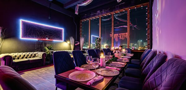 Лофт Овертон. Темное помещение с длинным столом, сервированным тарелками и бокалами, окруженным мягкими стульями. На стене неоновая декоративная панель. Большие окна в раме выходят на ночной город. Диван стоит у стены. Светодиодные гирлянды на потолке.