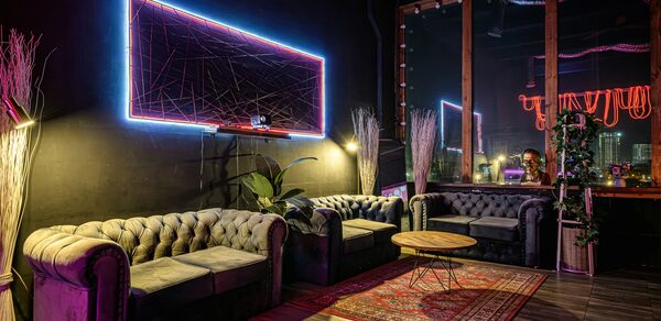 Лофт Овертон. Лофт-пространство с черными стенами и потолком, три серых дивана, низкий деревянный стол, ковёр с узором, неоновая подсветка на стене, большие окна, деревянный пол. Декор из растений в углах, стойка с зеркалом, стена с графическим узором в неоновом свете.