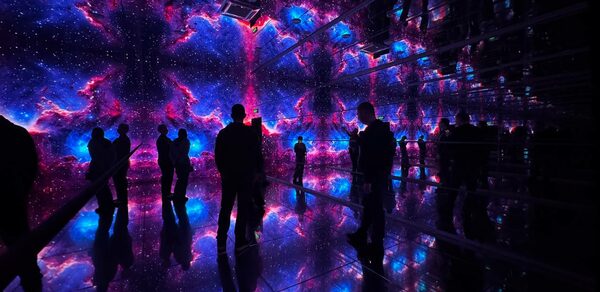 Futurione(Футурио́не). Интерактивное пространство с зеркальными стенами и полом, освещенное световыми проекциями космоса в ярких синих и фиолетовых тонах. Несколько человек стоят внутри, отражаясь в поверхностях.