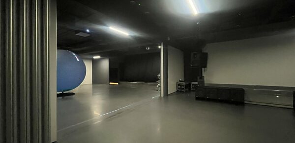 Арт-пространство до 600 гостей площадью 2000 м². Просторное помещение с серым полом и стенами, черный потолок с встраиваемым светодиодным освещением, синяя сфера у стены. Есть черные диваны, колонки, звуковое оборудование, металлическая конструкция с лампами.
