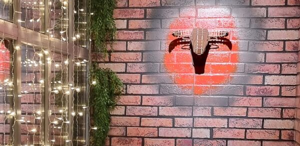 BULL LOFT(Лофт Бык) . Кирпичная стена с граффити в форме красного круга и черной гитары. Слева деревянная конструкция с висящими гирляндами и зеленью. Освещение теплое, мягкое.