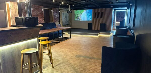 BULL LOFT(Лофт Бык) . Лофт-пространство с деревянными полами, барной стойкой, барными стульями, диванами, кирпичными стенами и экраном на стене. Современное освещение, тёмные потолки и декоративные панели.