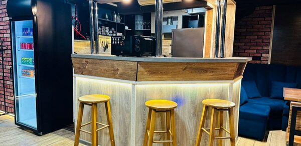 BULL LOFT(Лофт Бык) . Барная стойка с деревянной обивкой и тремя высокими стульями, подсветка снизу, фонарики сверху. В глубине shelving с бутылками и стаканами. Напольное покрытие светлое. В углу черный холодильник с напитками. Справа диван темного цвета.