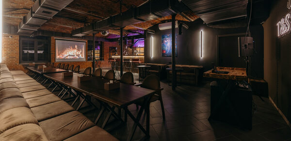 Авторские пространство T&S loft (ти энд си лофт) . Просторное помещение с кирпичными стенами и деревянным потолком, длинные деревянные столы и скамейки, игровые столы, проектор с изображением камина, настенные светильники, тёмные плиточные полы, зона с барной стойкой и неоновой подсветкой.